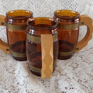 May include: A set of three ceramic mugs with a speckled, cream-colored glaze. Each mug features a different hand-painted floral design in shades of blue, green, and yellow. The mugs have a simple, rounded shape and a comfortable handle.