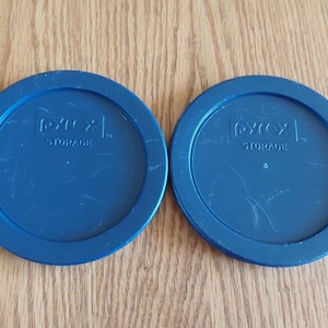 May include: Two blue Pyrex Storage lids. The lids are round with a slightly raised edge and the text "Pyrex STORAGE" embossed in the center. The lids are made of a durable, possibly plastic, material.
