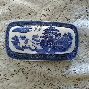 May include: A rectangular blue and white ceramic butter dish with a lid. The lid features a detailed blue and white Willow pattern scene. The dish sits on a matching base with a blue border.