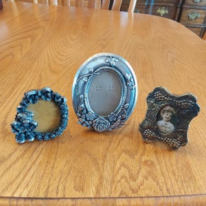 May include: Three silver-toned picture frames with ornate designs. The frame on the left has a teddy bear design, the middle frame has a floral design, and the frame on the right has a grapevine design.