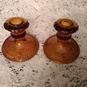 May include: A pair of amber glass candle holders with a decorative etched design. The holders have a round base and a fluted column.