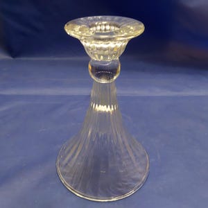 May include: Clear glass candlestick holder with a flared base and a round top. The candlestick has a bulbous midsection and a textured rim. The glass is transparent, allowing light to pass through. The candlestick is designed to hold a single candle.