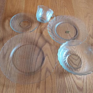 May include: A collection of clear glass tableware, including a teacup, a saucer, and three plates of varying sizes and designs. The plates have decorative edges and a heart-shaped design.
