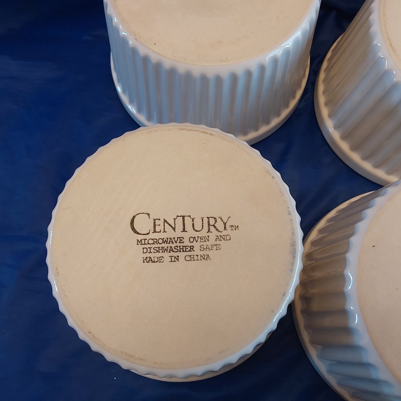 May include: Four white ceramic ramekins with a fluted design. The bottom of one ramekin displays the text "CENTURY" and "MICROWAVE OVEN AND DISHWASHER SAFE MADE IN CHINA". The ramekins are on a blue surface.