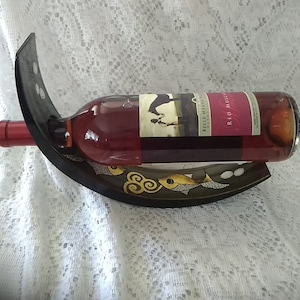 Puede incluir: Un porta botellas de vino de madera negra con un diseño curvo. El soporte presenta un diseño pintado en oro de peces y remolinos. Una botella de vino Belle Meade Red Muscadine está en el soporte.