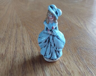 May include: A small porcelain figurine of a woman wearing a blue dress and hat. She is holding a bouquet of flowers.