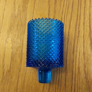 May include: A vibrant blue glass bottle stopper with a textured, diamond-patterned surface. The stopper has a rectangular shape with a small, cylindrical base. The object is set against a wooden background.