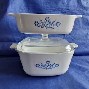 May include: Two white casserole dishes with blue floral designs. The top dish is rectangular, and the bottom dish is square with a clear glass lid. Both dishes have handles. The dishes are stacked on top of each other.