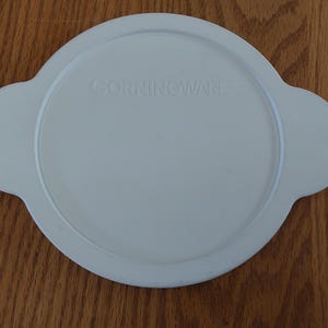 May include: A white CorningWare lid with two side handles. The lid is round with the brand name embossed in the center. The lid is on a wooden surface.