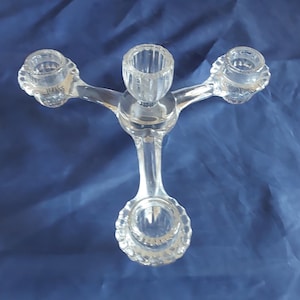 May include: Clear glass candelabra with four candle holders. The central holder is cylindrical, and the others are round. The candelabra has a decorative, branching design. The piece is made of clear glass and sits on a blue surface.