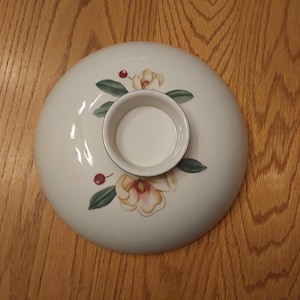 May include: A white ceramic serving dish with a central circular indentation. The dish is decorated with floral and berry motifs in shades of yellow, green, and red. The dish is set against a wooden surface.