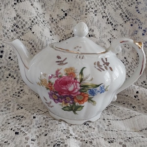 May include: White porcelain teapot with a floral design featuring pink, purple, orange, and blue flowers. The teapot has a gold trim and a lid with a small knob. The handle has a gold accent.