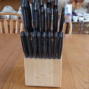 May include: A set of black knives with silver accents in a wooden block. The knives are arranged vertically in a light-colored wooden block. The block has the text "Paula Deen" on the front.