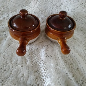 May include: Two brown and white ceramic soup bowls with lids and handles. The bowls are on a white lace tablecloth.