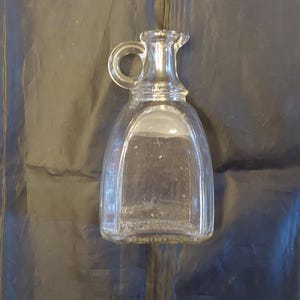 May include: A clear glass cruet with a handle and a pouring spout. The cruet has a rounded body and a small handle. The glass is transparent, allowing the contents to be visible. The cruet is likely used for serving oil or vinegar.