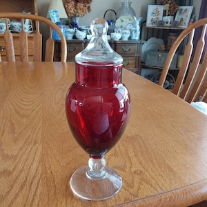 May include: A clear glass jar with a red glass body and a clear glass lid. The jar has a round base and a tall, slender body. The jar is sitting on a wooden table.