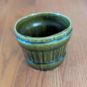 May include: A small, green ceramic planter with a ribbed design. The planter has a rounded shape with a blue accent band near the top and bottom. The interior is dark green.