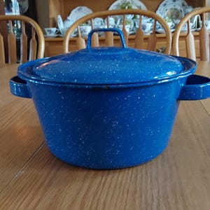 May include: A blue speckled enamelware pot with a lid and side handles. The pot has a handle on the lid for easy lifting. The pot is sitting on a wooden table. The pot is likely used for cooking.