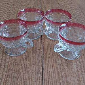 May include: Four clear glass dessert cups with a ruby red rim and handle. The cups have a diamond pattern and sit on a small footed base. The cups are arranged on a wooden surface.