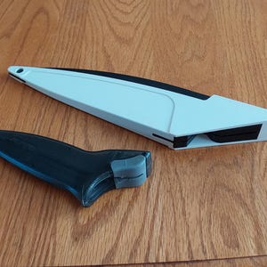 Op de afbeelding: Een wit en zwart keukengereedschap, mogelijk een messenslijper, ligt op een houten oppervlak. Het gereedschap heeft een lange, schuine witte behuizing met een zwart accent en een aparte zwarte handgreep met een grijze grip.
