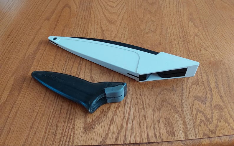 Op de afbeelding: Een wit en zwart keukengereedschap, mogelijk een messenslijper, ligt op een houten oppervlak. Het gereedschap heeft een lange, schuine witte behuizing met een zwart accent en een aparte zwarte handgreep met een grijze grip.