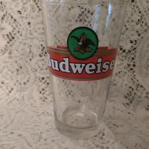 May include: A clear glass Budweiser beer glass with a red and gold Budweiser logo and a green circle with a stylized eagle.