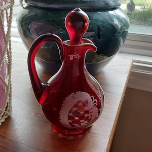 May include: A ruby red glass cruet with a stopper and handle. The cruet has a white etched design on the front. The cruet is sitting on a wooden surface. A pink container and a green vase are in the background.
