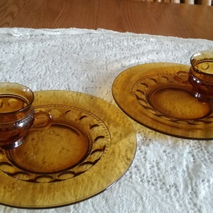 May include: Two amber glass dessert plates with matching cups. The plates have a raised rim with a scalloped design. The cups have a textured surface with a raised design.