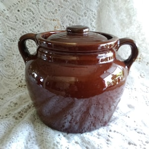 May include: A brown ceramic bean pot with a lid and two handles. The pot is glazed and has a shiny finish.