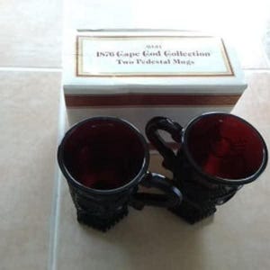 May include: Two dark red pedestal mugs with black handles and bases. The mugs are part of the "1876 Cape Cod Collection" and are displayed next to their original box. The box is white with a brown and gold border.