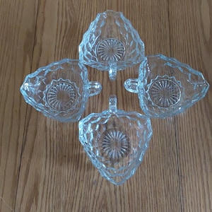 May include: Four clear glass serving dishes arranged in a cross shape. Each dish has a unique, leaf-like shape with a handle and a textured, circular design in the center. The dishes are on a light brown wooden surface.
