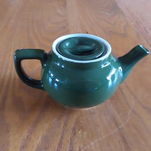 May include: A small, dark green ceramic teapot with a rounded body, a curved spout, and a handle. The lid is dark green with a white rim. The teapot is sitting on a wooden surface.
