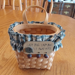 May include: A small woven basket with a blue and white checkered fabric trim. The basket has a wooden handle and a sign that reads "Lady Bug - Lady Bug Fly Away Home".