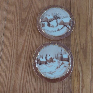 May include: Two oval-shaped porcelain plates with a gold trim. Each plate features a winter scene with brown and white houses and trees. The top plate has the text "Hollinsworth" in the bottom right corner. The bottom plate has the text "Hollinsworth" in the bottom left corner.