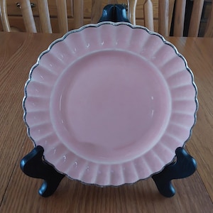 May include: A pink ceramic plate with a scalloped edge and a silver trim. The plate is on a black stand.