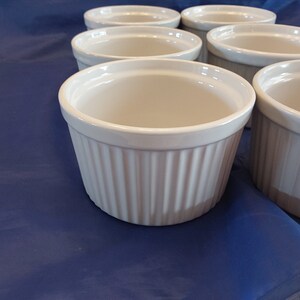 May include: A set of six white ceramic ramekins with a fluted exterior. The ramekins are round with a smooth interior and are arranged on a blue surface. These are ideal for baking individual portions of desserts or savory dishes.