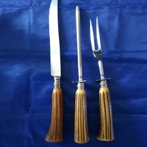 K&ouml;nnte beinhalten: Ein Set aus drei silberfarbenen K&uuml;chenutensilien mit braunen und gelben Hirschhorn-Griffen. Das Set enth&auml;lt ein Schnitzmesser, eine Schnitzgabel und einen Schleifstahl.