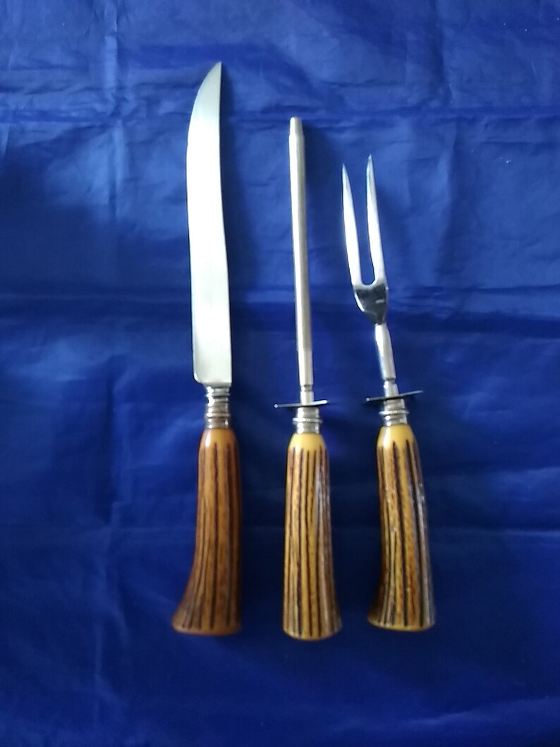 K&ouml;nnte beinhalten: Ein Set aus drei silberfarbenen K&uuml;chenutensilien mit braunen und gelben Hirschhorn-Griffen. Das Set enth&auml;lt ein Schnitzmesser, eine Schnitzgabel und einen Schleifstahl.