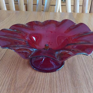 May include: A ruby red glass bowl with a ruffled edge and a pedestal base. The bowl has a glossy finish and a small, round glass accent in the center. The bowl is likely vintage and is suitable for decorative use.