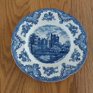 May include: A white and blue decorative plate with a scalloped edge. The plate features a blue landscape scene of a castle and floral patterns. The plate is set on a wooden surface.