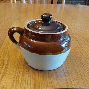 May include: A brown and white ceramic sugar bowl with a lid and handle. The bowl has a glossy finish and is sitting on a wooden table. The sugar bowl is a classic kitchenware item.