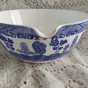 May include: A white ceramic bowl with a blue Willow pattern design. The bowl has a spout and a detailed landscape pattern around the rim. The bowl is sitting on a white lace tablecloth.