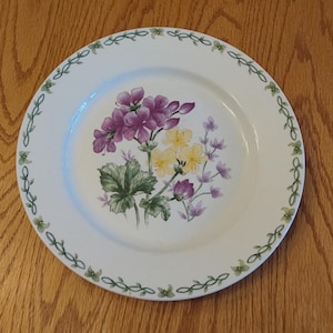 May include: White ceramic dinner plate with a green vine border and a floral design featuring purple, yellow, and green flowers.