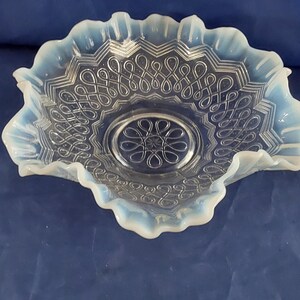 May include: A clear glass bowl with a ruffled edge and intricate swirl design. The bowl has a central circular design with a floral pattern. The glass has a slight opalescent quality, with a pale blue hue around the edges.