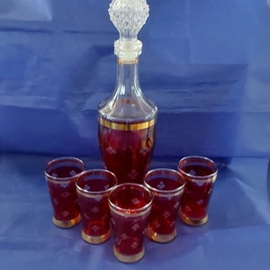 May include: A set of six red glass shot glasses with gold trim and a clear glass decanter with a crystal stopper. The glasses and decanter have a white floral pattern.