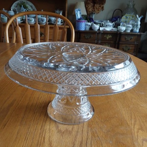 May include: A clear glass cake stand with a decorative, raised, floral design on the top and a diamond pattern around the edge. The stand has a pedestal base.