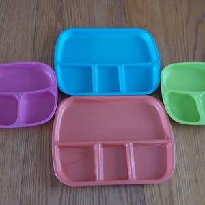 May include: Four divided plastic plates in different colors: purple, blue, orange, and green. Each plate has three sections.
