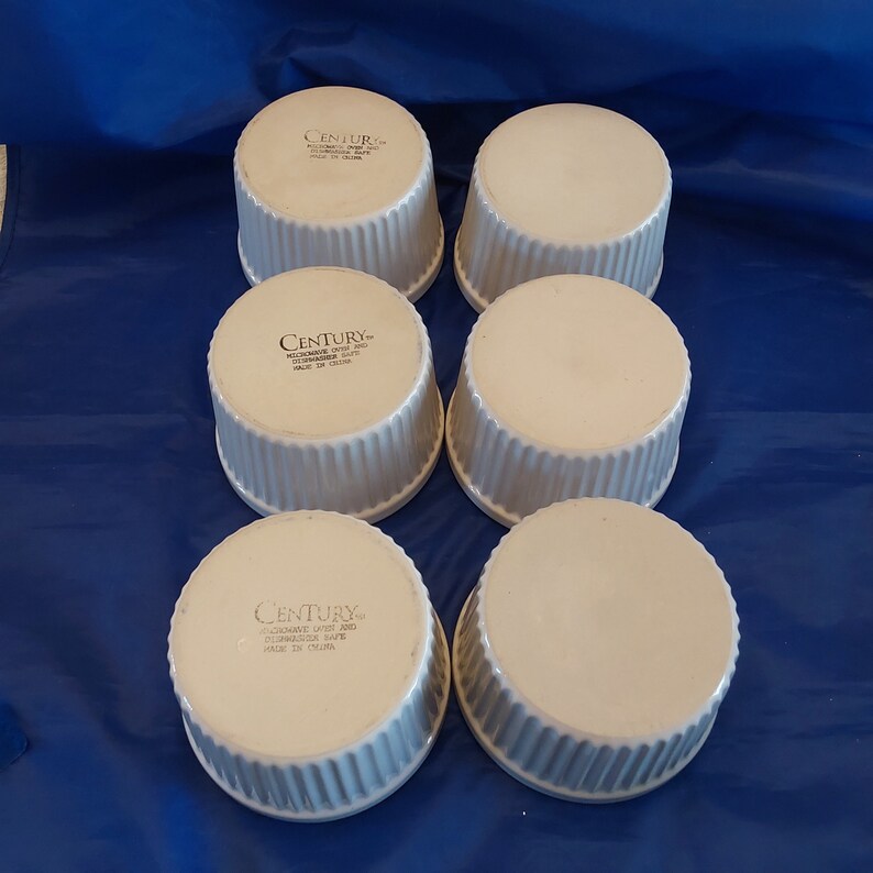 May include: Six white ceramic ramekins with a ribbed exterior and a smooth interior. The ramekins are arranged in two rows of three on a blue surface. Each ramekin has the word "CENTURY" printed on the bottom.