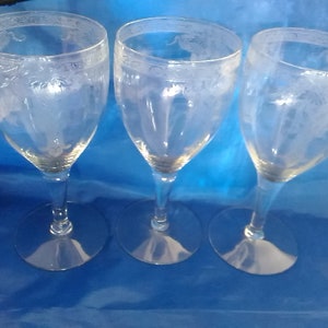 May include: Three clear glass wine glasses with etched floral designs. The glasses are arranged in a row on a blue surface.