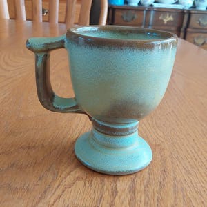 May include: A green and brown ceramic mug with a unique handle and a pedestal base.